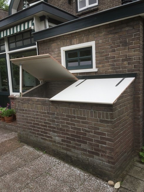 Gemetselde opbergkist met opengeklapte witte deksels tegen een bakstenen gevel